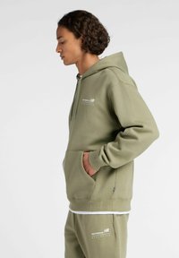 Olijf groene katoenen hoodie met een kangoeroezak, geribde manchetten en een capuchon met trekkoord. Bevat witte "New Balance" branding op de borst.