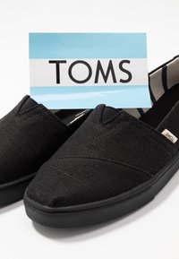 Svarta kanvas slip-on skor har en texturerad ovandel, elastisk insats och en gummisula. TOMS-logotypen visas på en blå etikett.