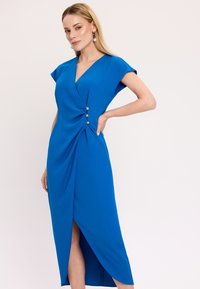 Kobaltowa sukienka z rękawami typu cap, głębokim dekoltem w serek, drapowanym fasonem i trzema ozdobnymi guzikami w talii. Spódnica ma boczne rozcięcie.