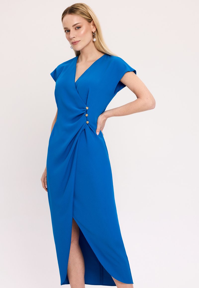 Kobaltowa sukienka z rękawami typu cap, głębokim dekoltem w serek, drapowanym fasonem i trzema ozdobnymi guzikami w talii. Spódnica ma boczne rozcięcie.
