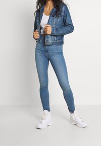 Chaqueta vaquera azul oscuro con dos bolsillos en el pecho, cierre de botones y vaqueros ajustados de talle alto en azul claro. Zapatillas blancas completan el conjunto.