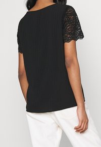 Haut noir en tissu côtelé, manches courtes en dentelle et encolure arrondie. L'ourlet est délicatement arrondi. Associé à un pantalon de couleur claire.