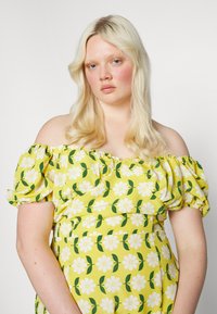 Vestido amarelo floral com mangas bufantes à descoberto. Apresenta flores brancas com folhas verdes e um design ajustado, feito de um tecido leve.