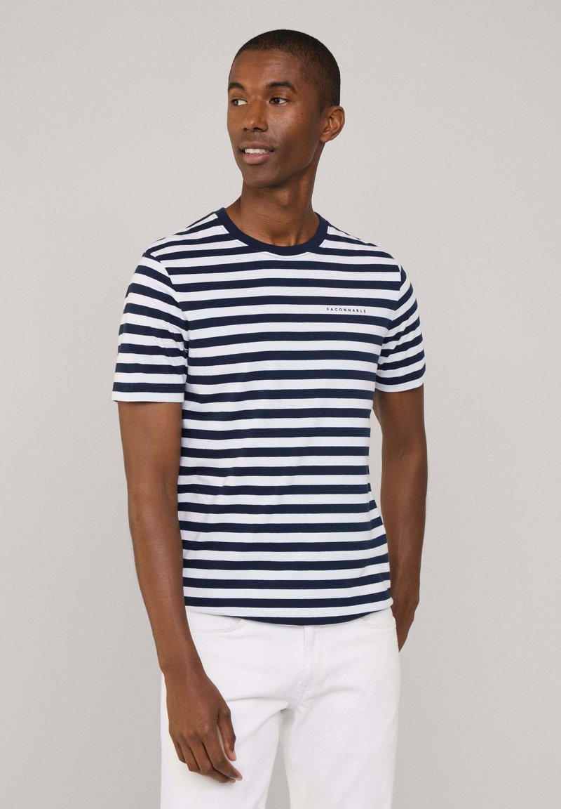 Granatowo-biała, paskowana koszulka z krótkim rękawem i okrągłym dekoltem, z małym logo na piersi, noszona z białymi spodniami.