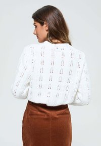 Pull en tricot blanc avec un motif ajouré texturé et un col arrondi, associé à une jupe en velours côtelé marron.