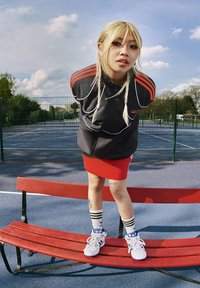 Crna zip-up jakna s crvenim trakama, crvena uska suknja, bijele tenisice s plavim akcentima, stoji na crvenoj klupi, vanjski sportski teren.