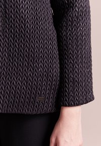 Prošívaná černá bunda s vzorem ve tvaru šípu, hladkým povrchem a viditelným logem u spodního lemu. Rukávy do lokte.