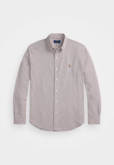 Krekliņš ar pogām, izgatavots no kokvilnas, ar šaurām vertikālām svītrām tumši sarkanā un krēmkrāsā. Tam ir klasiskā apkakle un neliela logo uz krūtīm.