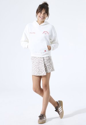 Sudadera blanca con capucha y texto rojo, combinada con una falda pantalón beige con estampado de leopardo. La modelo lleva zapatillas marrón claro con rayas blancas.