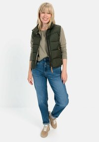 Grüne gepolsterte Weste mit Reißverschluss über einem hellgrünen Oberteil, kombiniert mit hellblauen Jeans und beigen Sneakern. Glatte Textur, lässige Passform.