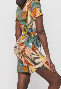 Femme portant une robe courte colorée et à motifs avec une ceinture, posant avec une main sur sa cuisse devant un fond uni.