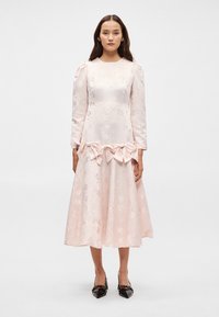 Φ�όρεμα ροζ με μακριά μανίκια και λουλουδάτο μοτίβο, πλισέ μέση με λεπτομέρειες σε σχήμα φιόγκου, φουσκωτή φούστα και λεία σατέν υφή. Μαύρες μυτερές γόβες.