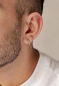 Orecchino a cerchi argento mostrato sul lobo dell'orecchio di un uomo, con una superficie liscia e lucida e un design circolare. Texture della pelle e capelli visibili.