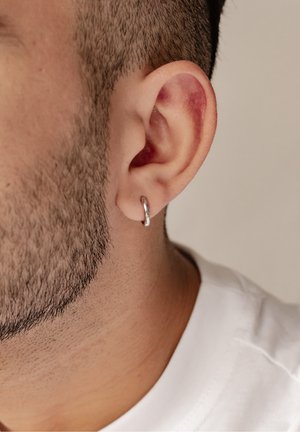 Pendiente de aro plateado mostrado en el lóbulo de la oreja de un hombre, con una superficie lisa y brillante y un diseño circular. Se puede ver la textura de la piel y el cabello.