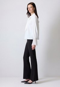 Blusa blanca con detalle de lazo y puños con pliegues, combinada con pantalones negros de campana y tacones de punta en un fondo neutro.
