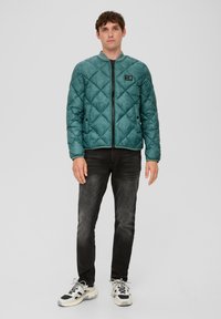 Gequiltete türkisfarbene Jacke mit Stehkragen und Reißverschluss, kombiniert mit dunklen Jeans und mehrfarbigen Sneakern.
