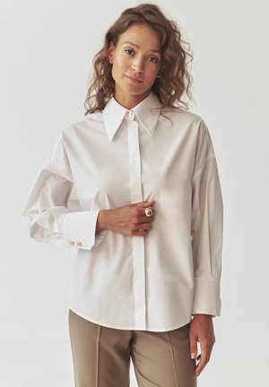White button-up shirt with a large collar, long sleeves, and gold-button accents. Smooth cotton fabric; relaxed fit, paired with beige trousers.