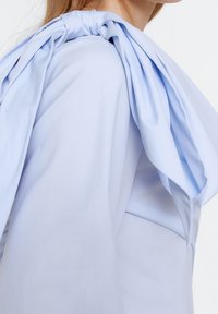 Vaaleansininen paita, jossa on suuri, solmittu nauha olkapäässä. Kangas on sileä, pehmeällä taipumisella ja valuvalla muotoilulla.
