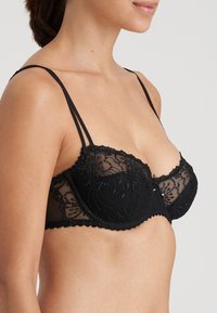 Svart spets-bh med genomskinliga kupor och blommönster, justerbara tunna axelband och en vågig kant. Inkluderar dekorativt centralt detalj.
