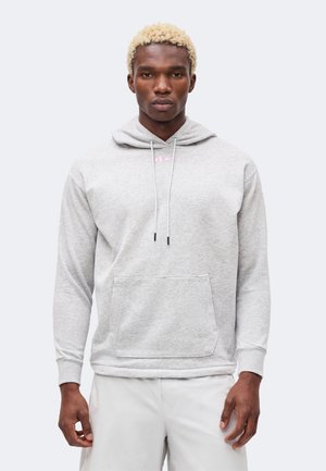 Hombre con cabello rubio que lleva una sudadera con capucha gris claro con logo rosa y pantalones blancos, de pie contra un fondo liso y claro.