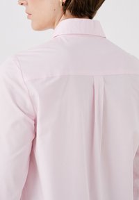 Camisa rosa clara con un elegante cuello, un pliegue en la parte posterior para mayor libertad de movimiento y una textura de tela suave.