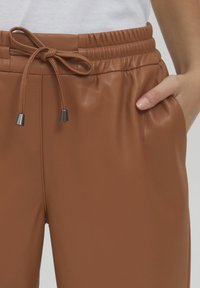 Brune faux læder joggers med elastisk talje, snøre lukning og sidelomme. Glat tekstur og tapered design.