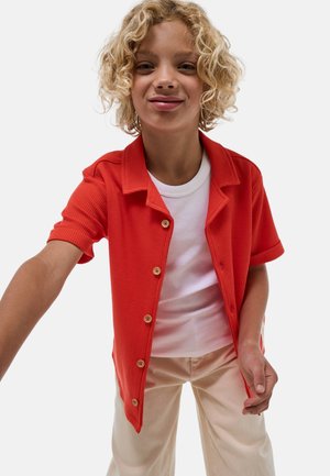 Chemise orange à manches courtes avec un tissu texturé, dotée d'un col pointu et de boutons en bois, portée par-dessus un débardeur blanc avec un pantalon couleur crème.