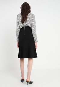 Svart utsvängd kjol med en åtsittande midja och dragkedjestängning, kombinerad med en blus med svart-vit zigzag-mönster, bärs med svarta klackar.