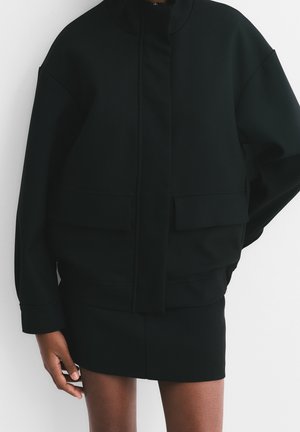 Personne portant une veste noire surdimensionnée avec des poches avant et une mini-jupe noire, debout devant un fond blanc uni.