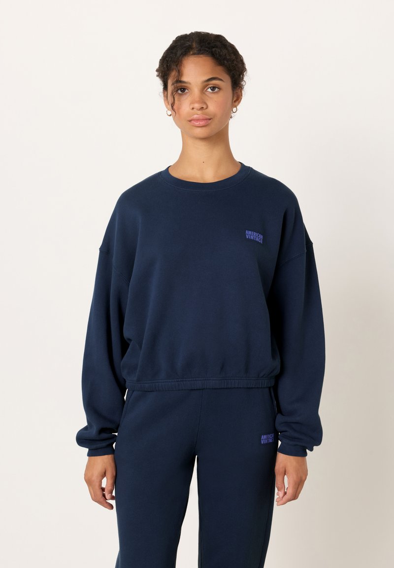 Tamsiai mėlynas džemperis iš minkštos medžiagos su nuleistais pečiais, dryžuotais rankogaliais ir apačia. Ant krūtinės mažas violetinis „American Vintage“ logotipas.