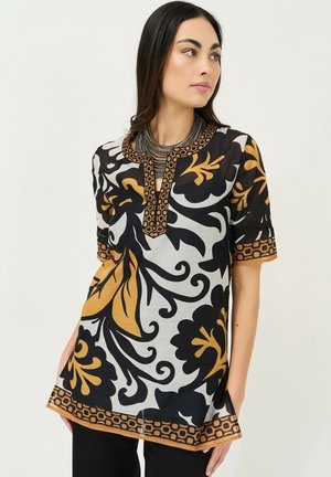 Femme portant une tunique à motif floral noir, blanc et moutarde avec une bordure géométrique, associée à un collier métallique superposé et un pantalon noir.
