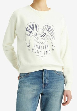 Osoba ubrana w białą bluzę Levi Strauss z logo orła i napisem "Quality Clothing San Francisco Cal", w zestawieniu z niebieskimi dżinsami.