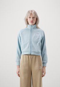 Veste zippée bleu clair avec un col montant et des poignets côtelés, arborant un petit logo Puma blanc sur la poitrine. Portée avec un pantalon de couleur or.