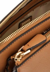 Pochette de texture tan avec des fermetures éclair dorées et des accents noirs. Présente plusieurs compartiments et un logo en relief sur la doublure intérieure.