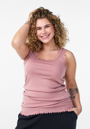 Nasmejana žena kovrčave kose nosi ružičasti top s čipkom, jednom rukom dira kosu, a drugom je u džepu, pokazujući tetovažu ruže na ruci.