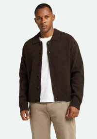 Cardigan de lana marrón con cuello tipo solapa, cierre frontal de botones, puños y dobladillo acanalados, combinado con una camisa blanca y pantalones beige.