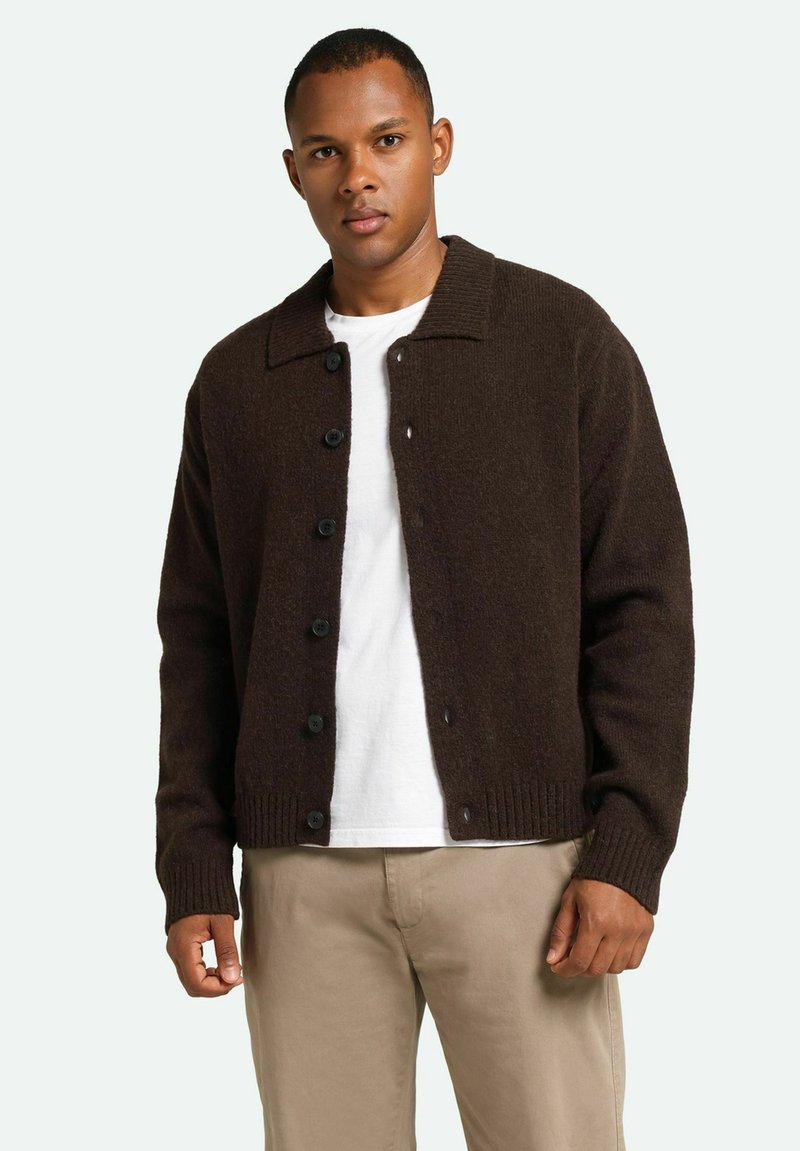 Cardigan de lana marrón con cuello tipo solapa, cierre frontal de botones, puños y dobladillo acanalados, combinado con una camisa blanca y pantalones beige.
