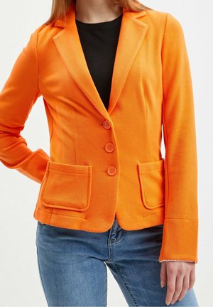Vrouw draagt een feloranje getailleerde blazer over een zwart topje met blauwe spijkerbroek, toont torso en een deel van een arm.