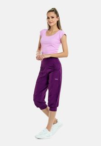Pantaloni viola fino al ginocchio con arricciature laterali, abbinati a una maglietta aderente rosa a maniche corte e scarpe da ginnastica bianche. Tessuto morbido e elasticizzato.