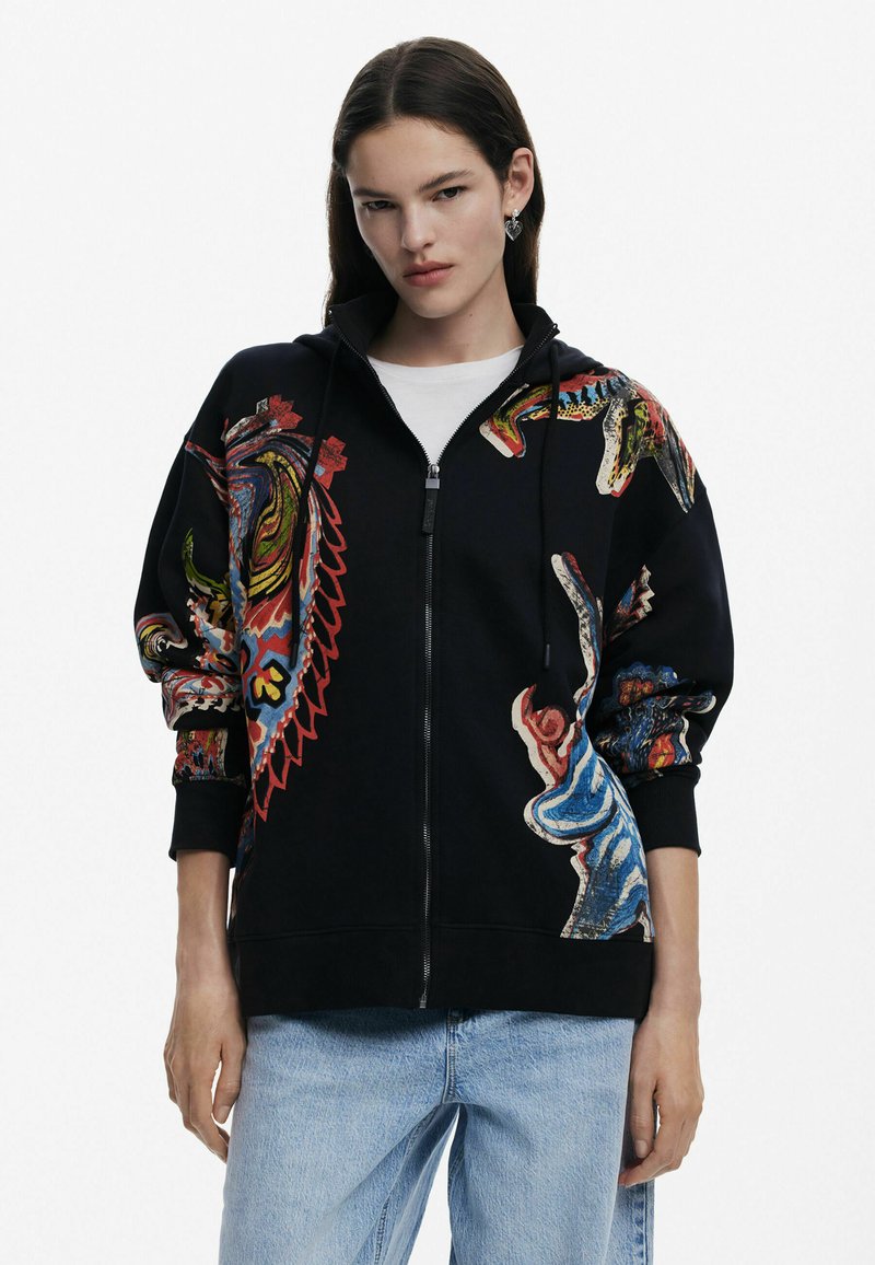 Svart zip-up hoodie med färgglada flerfärgsdesign, med en mjuk textur och ribbade muddar. Bärs över en vit skjorta och blå jeans.