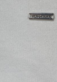 Tessuto bianco con una superficie liscia, caratterizzato da un'etichetta in metallo argentato incisa con la scritta "LASCANA". Lo sfondo è semplice e poco invadente.