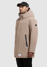 Giacca isolante beige con colletto alto, tasca con zip nera e patch con logo sulla manica. Presenta un tessuto morbido e una vestibilità rilassata.