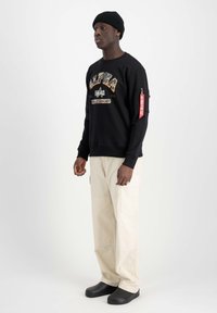 Felpa nera con il logo "ALPHA" colorato, pantaloni cargo beige, berretto nero e scarpe slip-on nere, con una tasca laterale con etichetta.