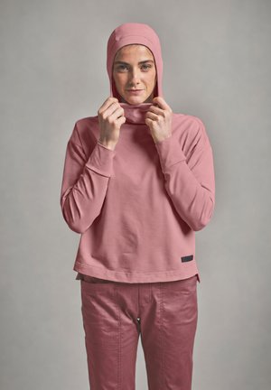 Woman facing forward adjusting a pink hooded top, wearing matching pink pants, against a plain gray background.