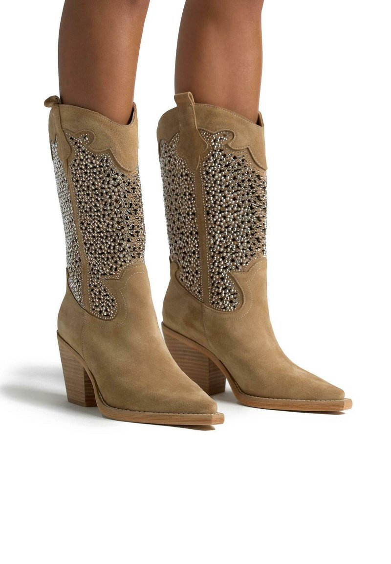 Botas de vaquero de ante marrón con punteras afiladas, adornadas con detalles de cuentas plateadas, presentan un tacón alto de bloque y recortes decorativos.