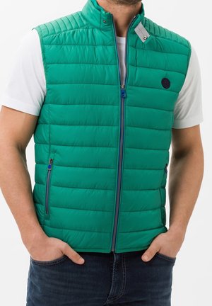 Homme portant un gilet matelassé vert vif sur une chemise blanche à manches courtes, mains dans les poches d'un jean foncé, sur un fond uni.