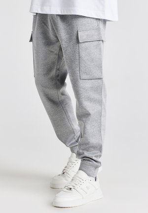 Personne portant un pantalon de survêtement cargo gris avec des poches latérales et des baskets blanches, debout sur un fond clair uni.