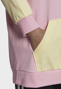 Felpa colorata con corpo rosa, maniche gialle, polsini a coste e una tasca frontale. Tessuto morbido con una consistenza liscia.