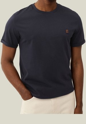 Mann trägt ein dunkelblaues T-Shirt mit kurzen Ärmeln und hochgekrempelten Bündchen sowie ein kleines oranges "II" Logo auf der linken Brust, kombiniert mit hellbeigen Hosen.