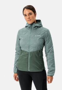 Giacca isolata verde con cappuccio, caratterizzata da un motivo a trapezio trapuntato nella parte superiore e una parte inferiore unita. Dotata di tasche con cerniera.